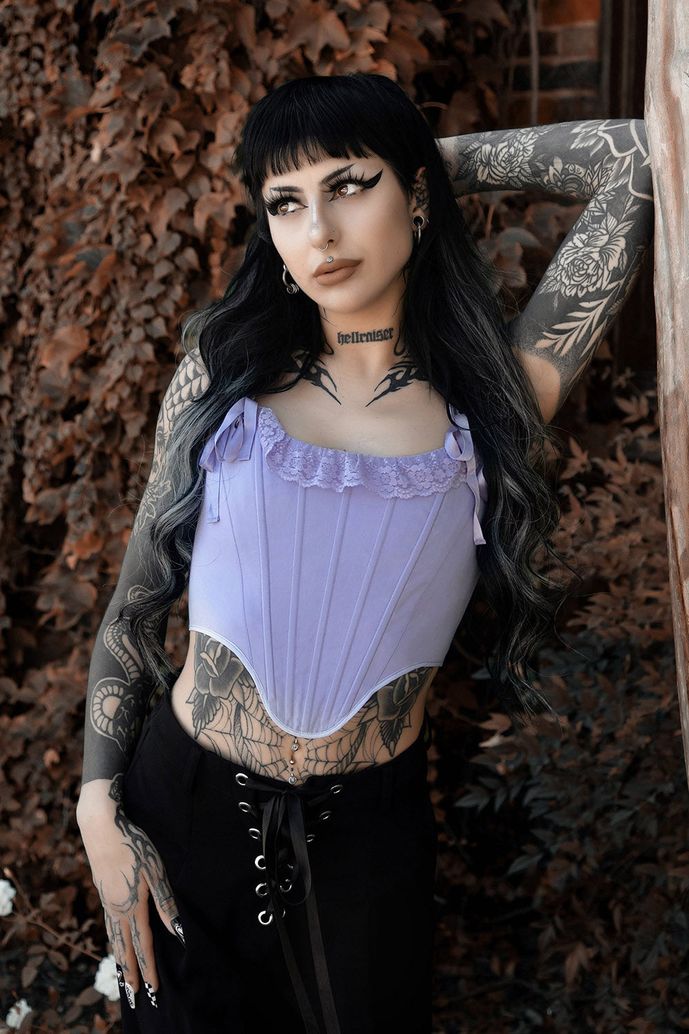 Person wearing a purple corset top with tattoos on arms and chest, standing against a textured wall.