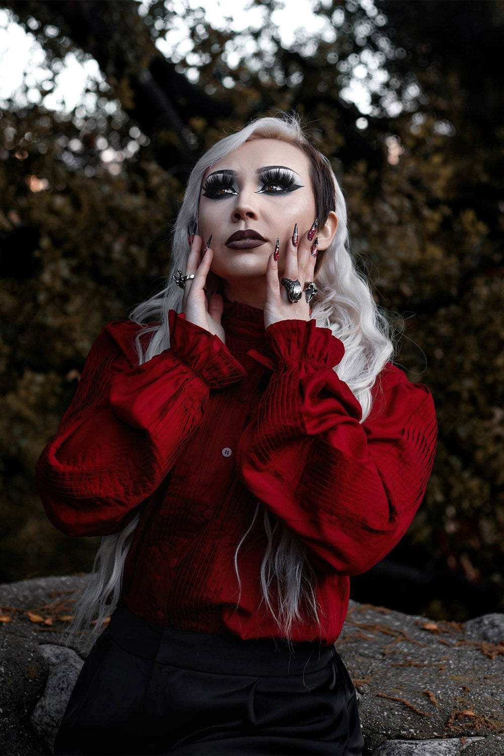 Person with white hair and dark makeup wearing a red blouse in a forest setting