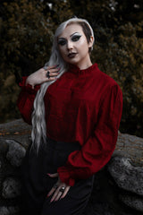 Person wearing a red blouse with dark hair and makeup, standing against a blurred natural background.