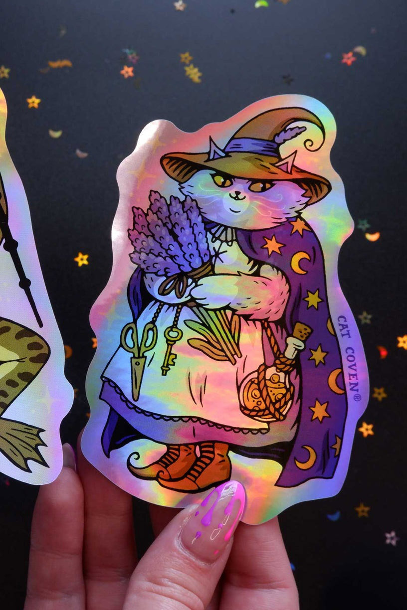 Holographic sticker of a cat in witch attire held by a hand against a dark background with star decorations.