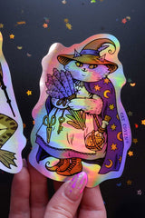 Holographic sticker of a cat in witch attire held by a hand against a dark background with star decorations.
