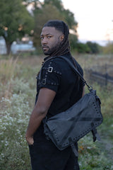 Dark punk messenger bag with distressed grey finish metal studs and utility compartments for industrial goth outfits and rebellious streetwear