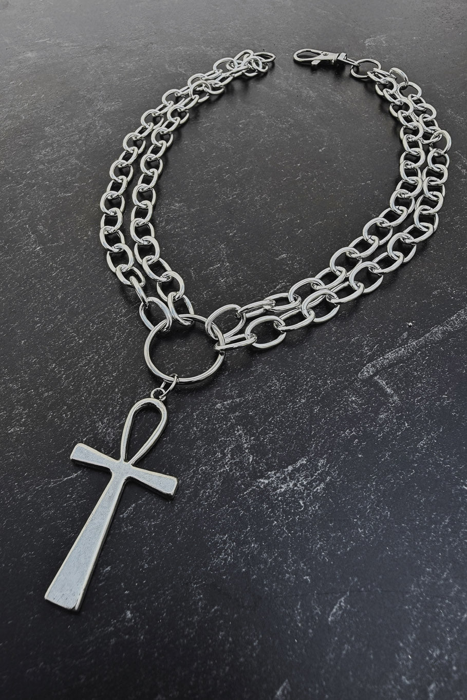 A silver-toned chain necklace featuring a double-layered curb chain, a central ring, and a dangling cross pendant, displayed on a dark textured slate surface.