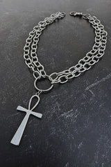 A silver-toned chain necklace featuring a double-layered curb chain, a central ring, and a dangling cross pendant, displayed on a dark textured slate surface.
