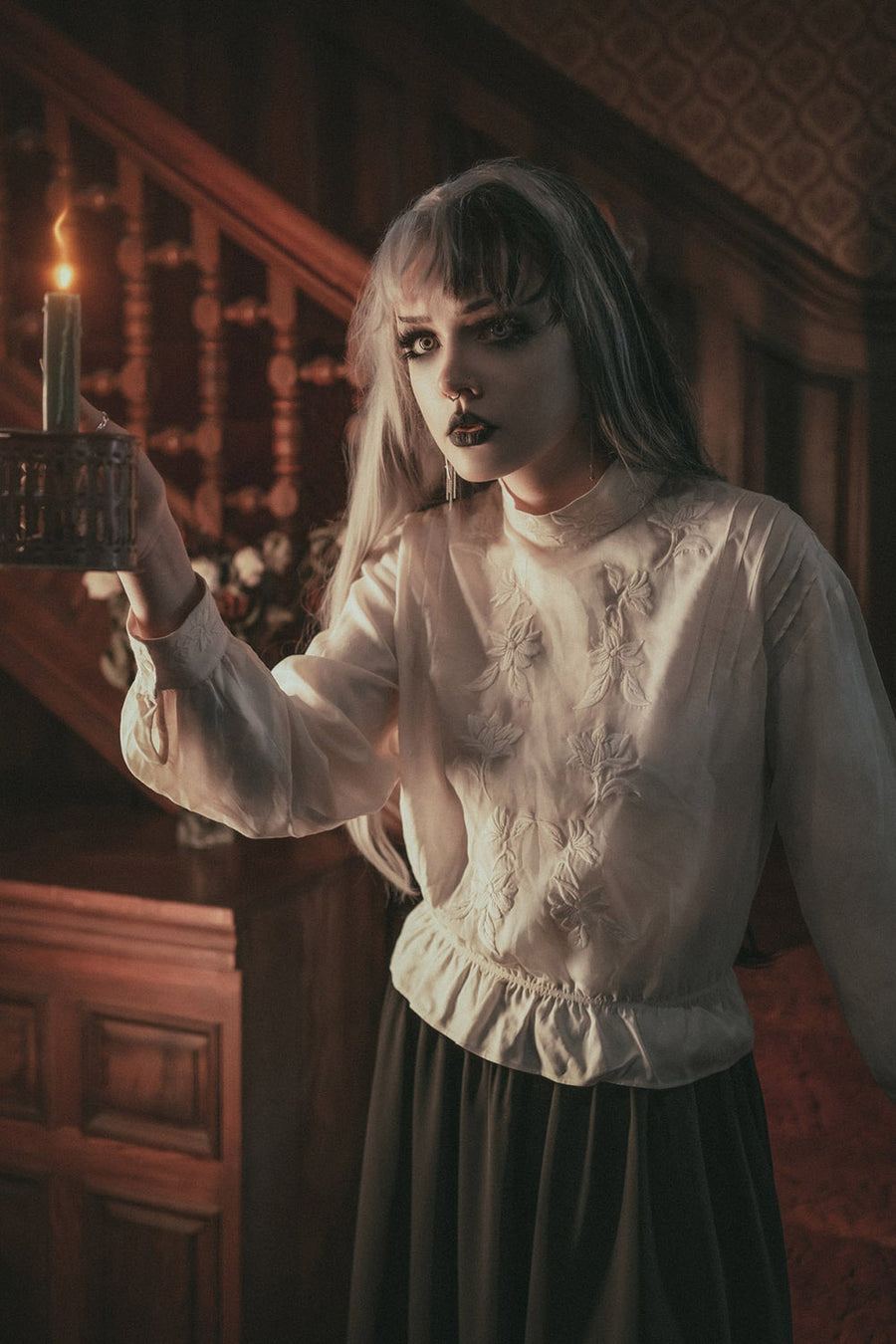 Woman in a white blouse and dark skirt holding a candle in a dimly lit room.