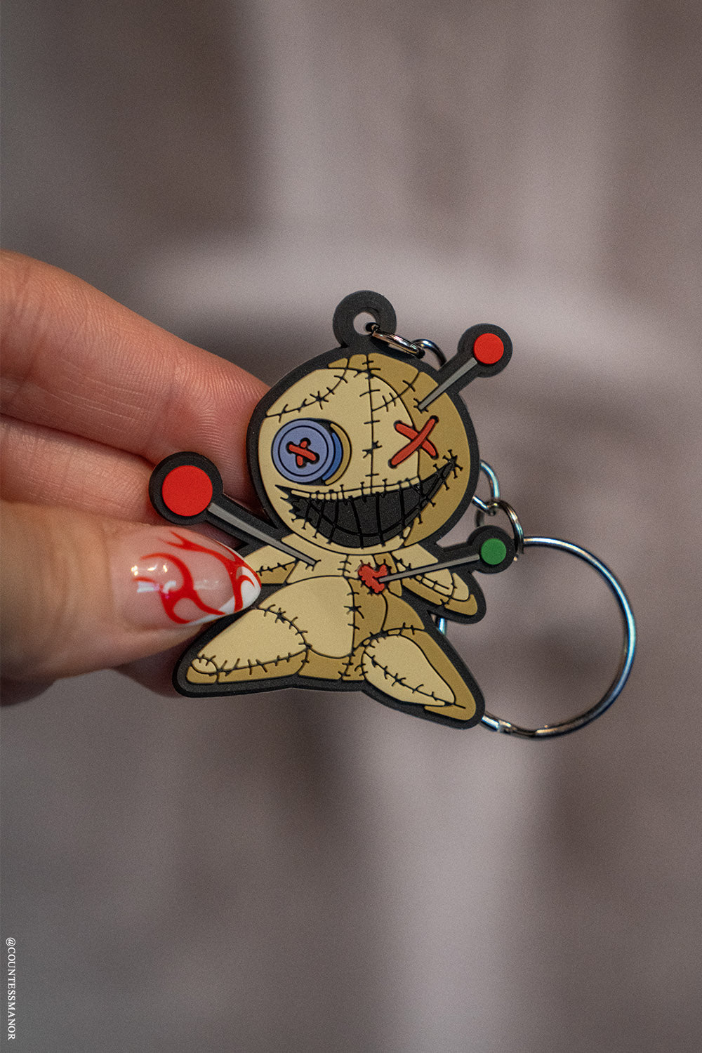 Voodoo doll keychain held by a hand with red nail polish against a blurred background