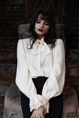 Woman wearing a white vintage goth inspired blouse with long sleeves and a high collar, sitting against a stone wall.