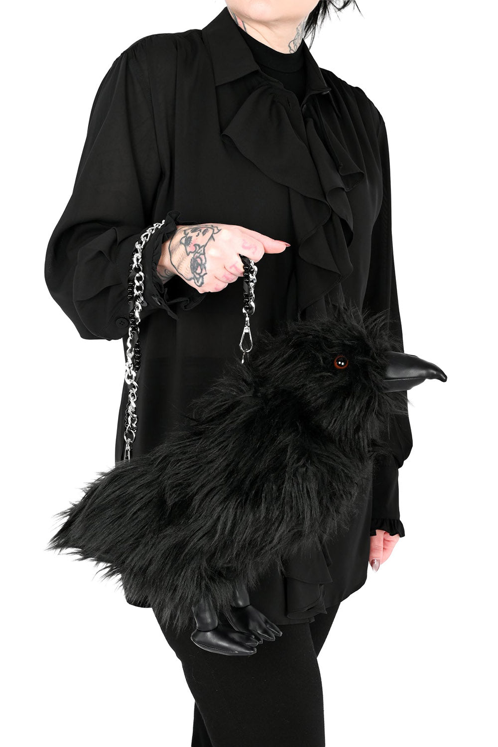 A person wearing a black ruffled blouse and black pants holds a fluffy black raven-shaped handbag with a silver chain strap against a plain white background.