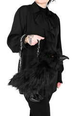 A person wearing a black ruffled blouse and black pants holds a fluffy black raven-shaped handbag with a silver chain strap against a plain white background.