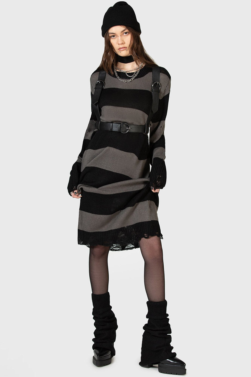 goth girl wearing a black and grunge gray striped dress with a high-waisted punk belted harness, black boots, and a black beanie on a white background.
