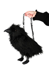 A black raven-shaped fluffy purse with a silver chain strap, held by a hand against a solid white background.