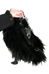 A black faux fur crow raven shoulder bag held by a hand wearing a black ruffled sleeve against a white background.