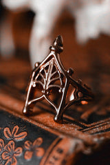 A close-up of a gothic-style spiderweb ring in a dark bronze metal, resting on a textured brown leather surface with a blurred background.