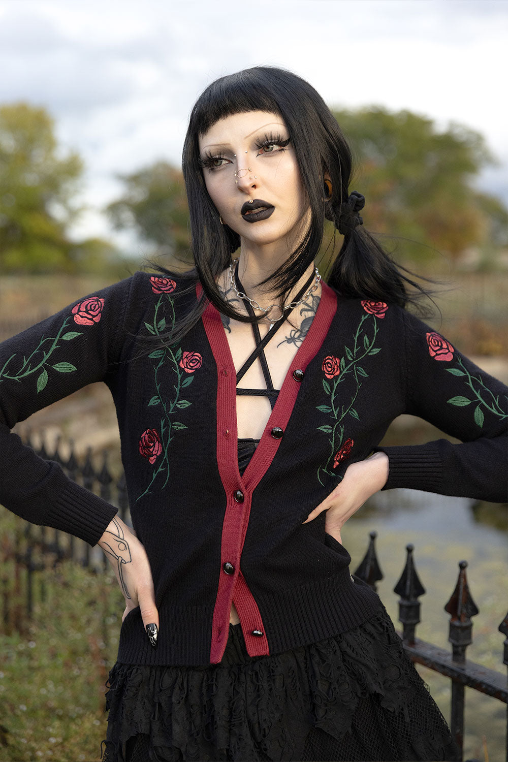 A gothic-style model wearing a black cardigan with red trim and red rose embroidery, posed outdoors in front of a fence and trees.