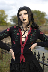 A gothic-style model wearing a black cardigan with red trim and red rose embroidery, posed outdoors in front of a fence and trees.