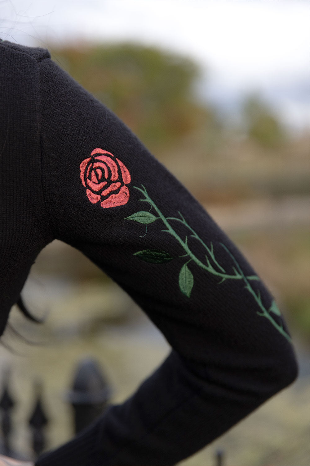 A close-up of a person's arm wearing a black long-sleeve top with an embroidered pink rose and green stem design on the upper sleeve, set against a blurred outdoor background.