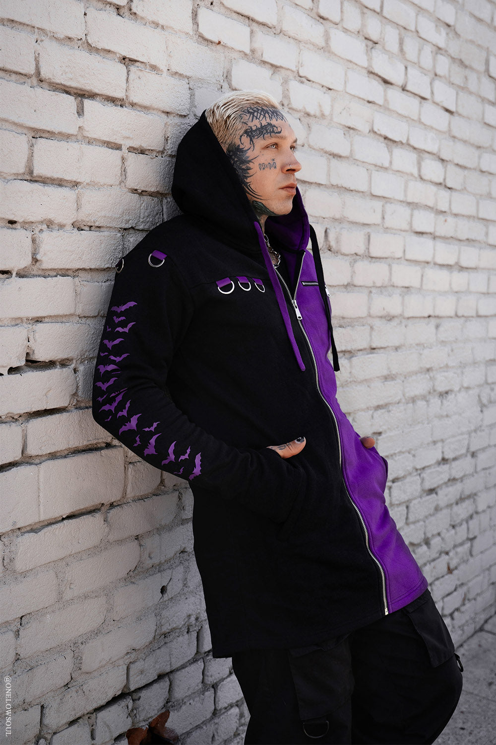 Person wearing a black and purple jacket leaning against a white brick wall.