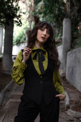 Woman in a green blouse and black vest standing outdoors with trees in the background