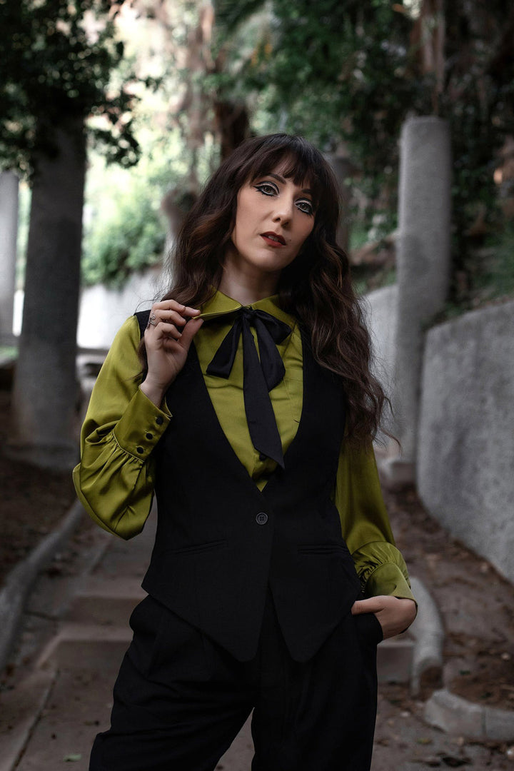 Woman in a green blouse and black vest standing outdoors with trees in the background