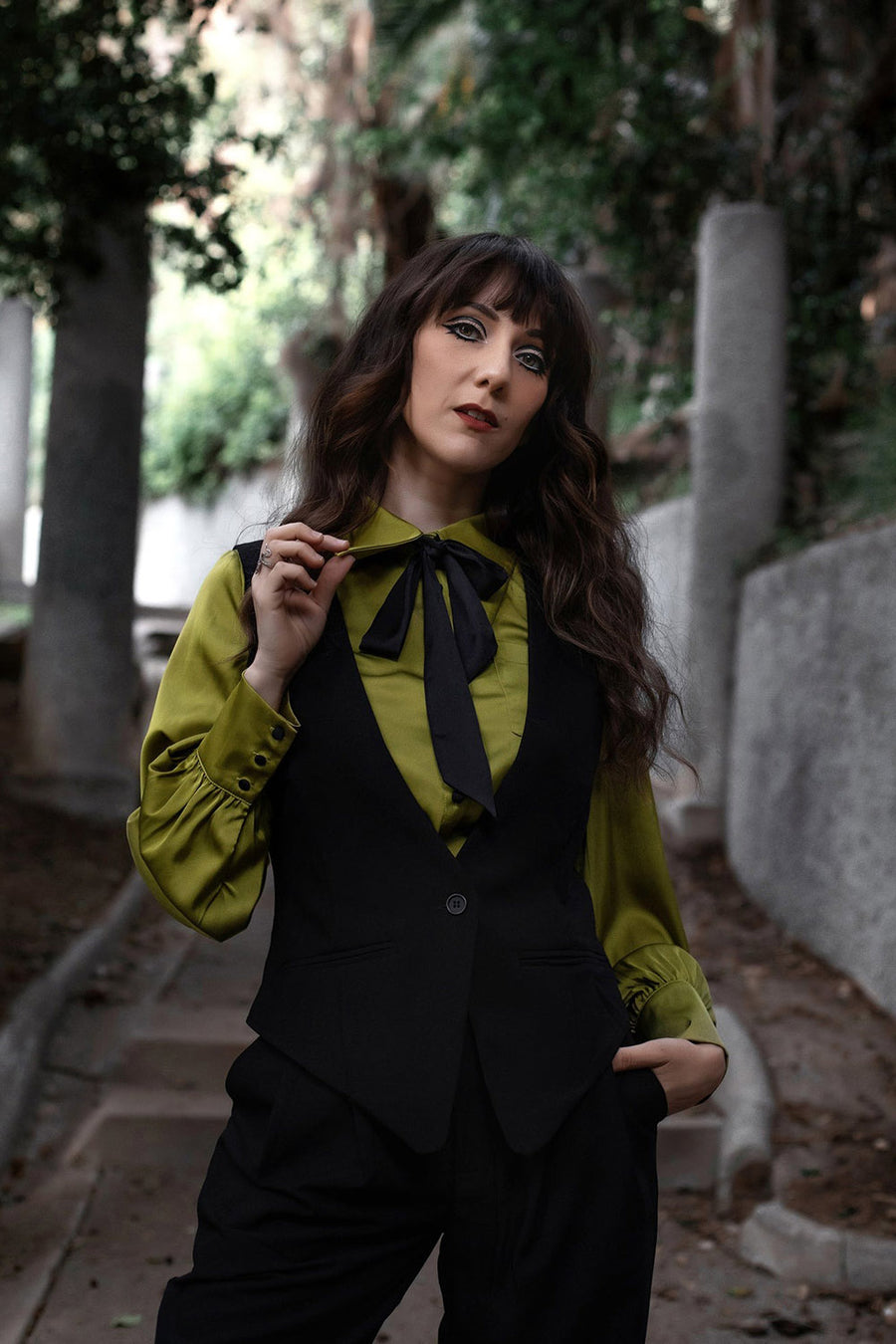 Woman in a green blouse and black vest standing outdoors with trees in the background