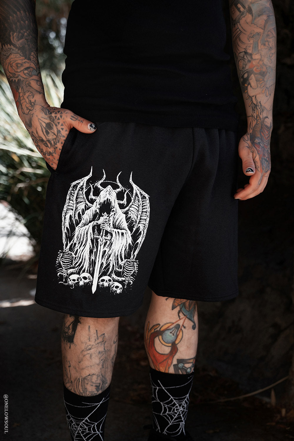 tattooed Person wearing black goth shorts with a white graphic design, standing outdoors.