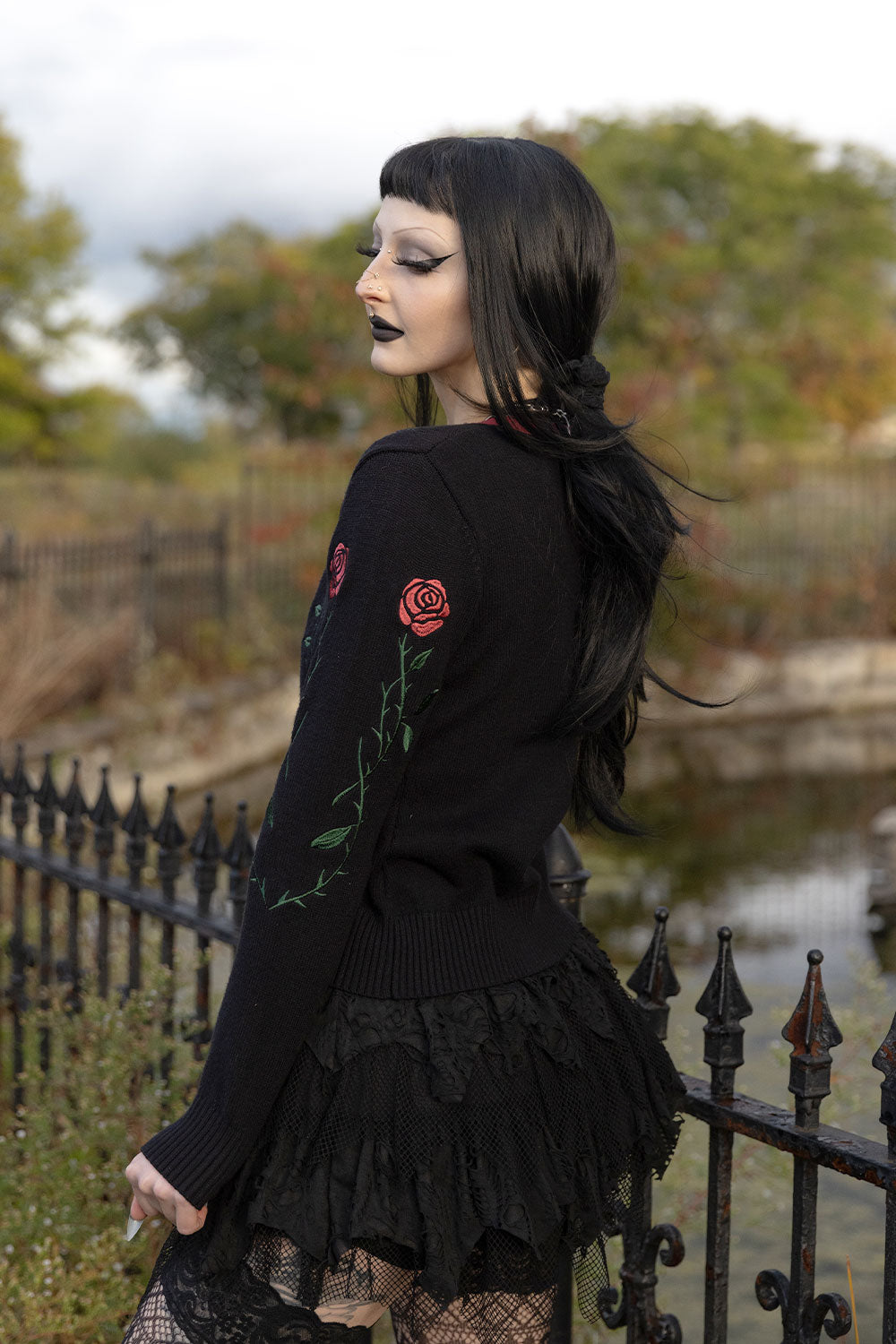 A woman with dark hair and gothic makeup is shown from the back wearing a black long-sleeve sweater decorated with red rose embroidery and a black ruffled skirt, standing outdoors near a fence and water.