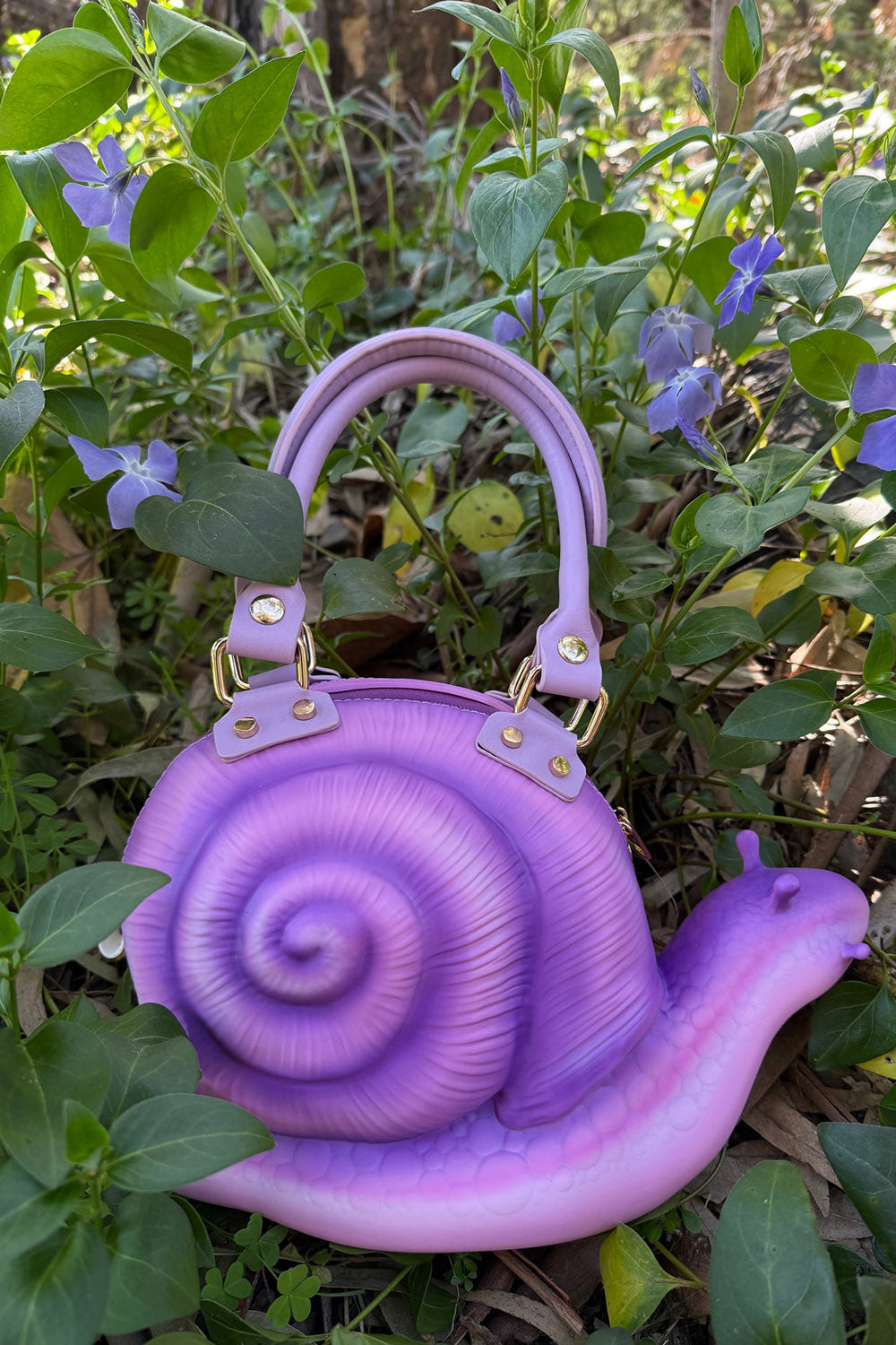 Purple snail-shaped handbag in a natural setting with green plants and purple flowers.