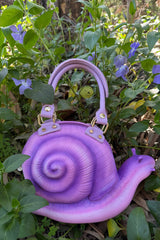 Purple snail-shaped handbag in a natural setting with green plants and purple flowers.