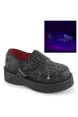 Black platform slip-on shoe with spiderweb embroidery, skeleton hand detail, and a small silver spider charm, shown on a white background with an inset demonstrating glow-in-the-dark effect under purple light.