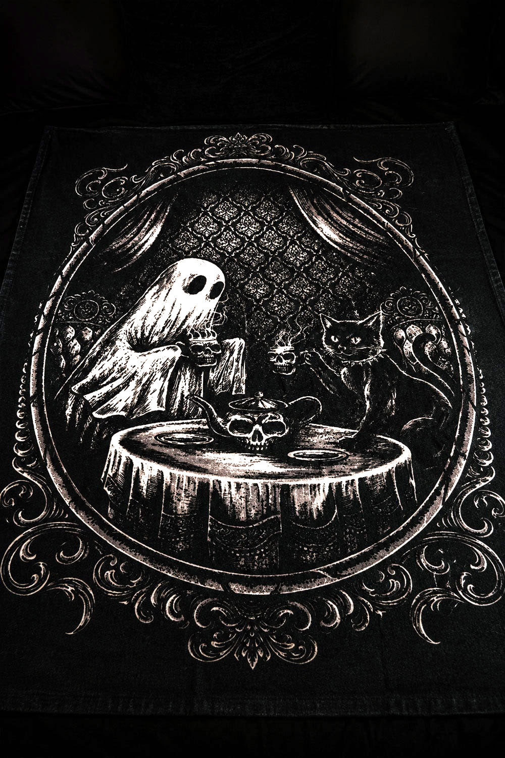 Gothic-style blanket with a ghost, cat, and skull teapot on a table within an ornate frame.