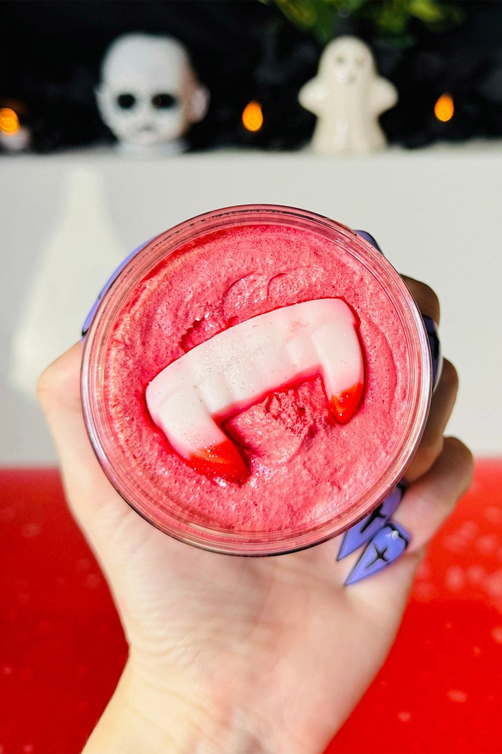Hand holding a red sugar scrub jar with a white vampire fang soap bar inside, with a Halloween-themed background