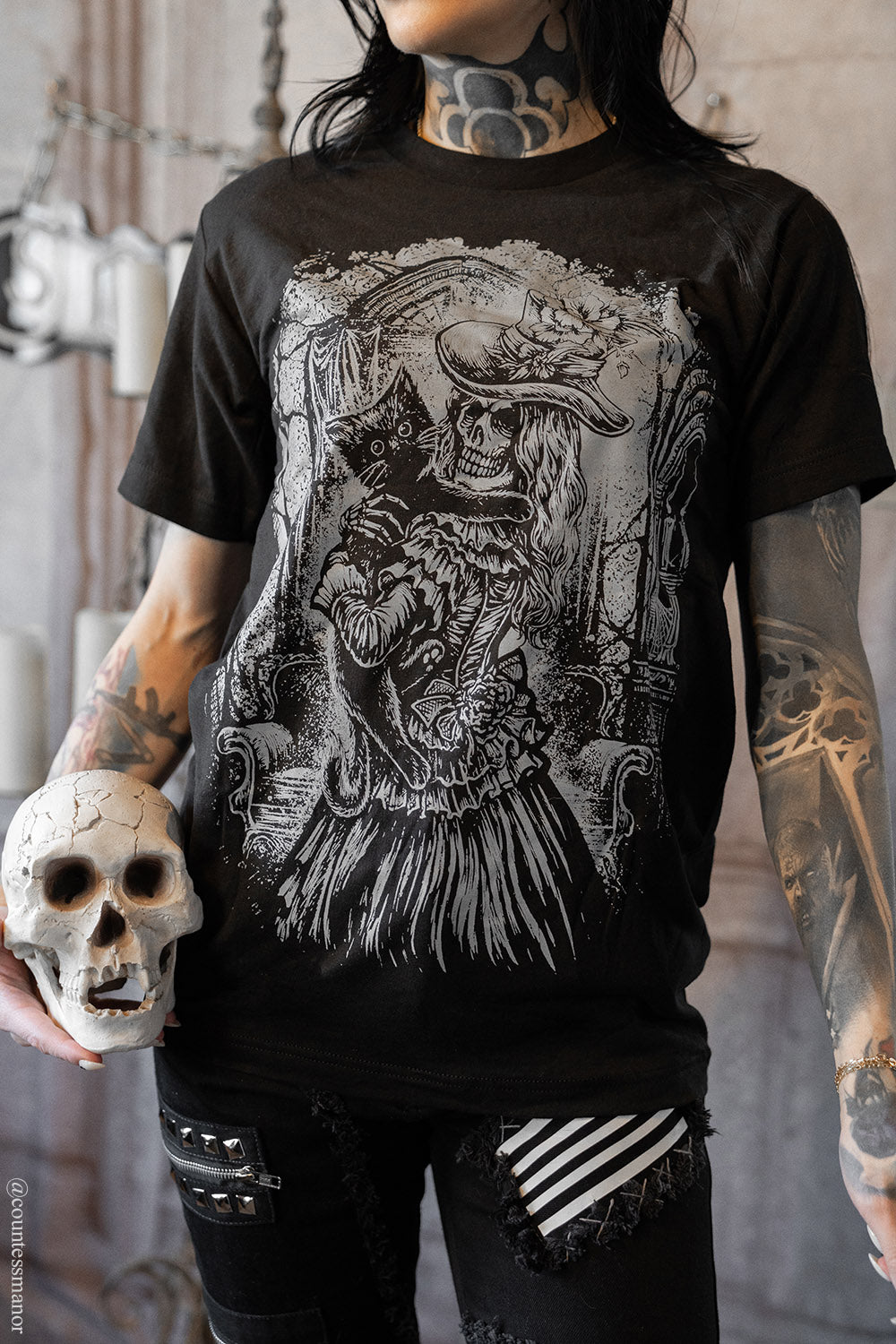 Person wearing a black t-shirt with a detailed graphic design of a victorian skeleton woman and a black cat holding a skull.