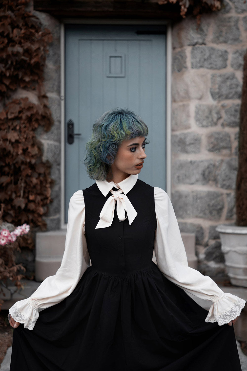 Person wearing a black dress with a lively ghosts white shirt and bow tie in front of a stone building.