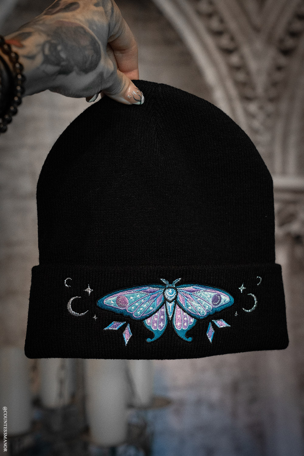 Black beanie with a colorful moth design held by a person with tattoos.
