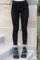 Black skinny pants with silver eyelet lace-up detailing down the sides, shown on a model standing on stone steps.