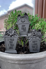 Three black tombstone-shaped garden stakes with humorous sayings in a pot.