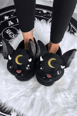 A pair of black cat slippers designed to resemble a character with large ears and a moon detail, worn by a person against a white furry background.