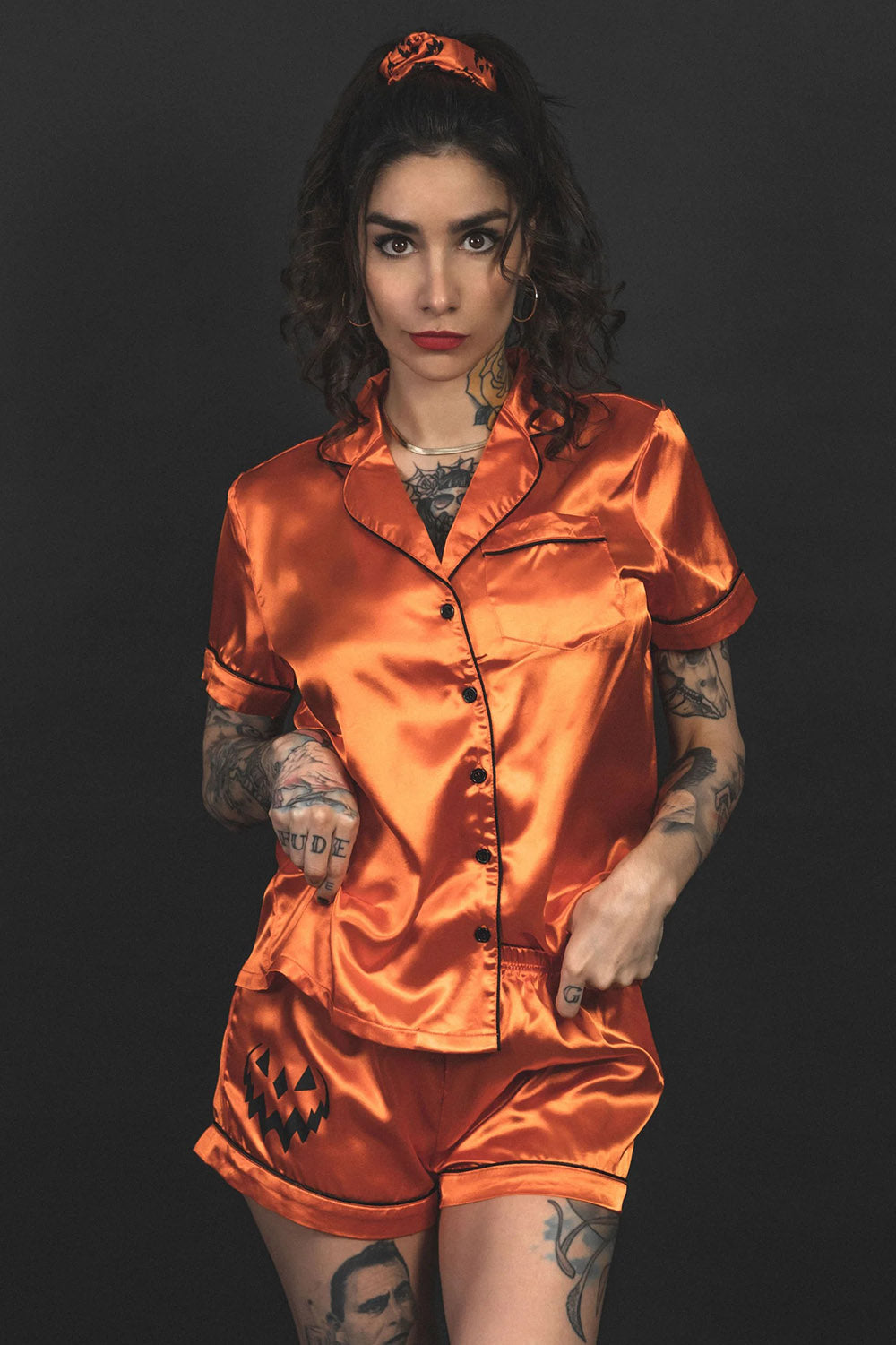 A person wearing an orange satin pajama set with button-up bodice and collar, featuring black contrast piping and sewn cuffs. The set includes a top, shorts, a hair scrunchie, and a sleep mask.